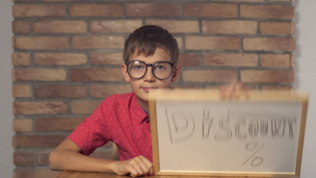child-sitting-at-the-desk-holding-flipchart-with-lettering-discount-on-the-background-red-brick-wall