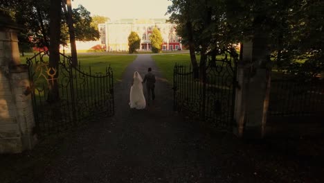 Aerial-drone-shot-of-a-groom-and-bride-moving-towards-mansion-in-a-park.