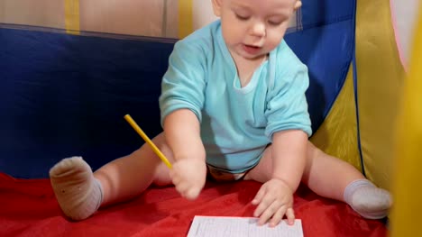 Attractive-baby-playing-in-the-children's-colorful-tent-in-the-house.-The-boy-carefully-draws-in-pencil-notebook