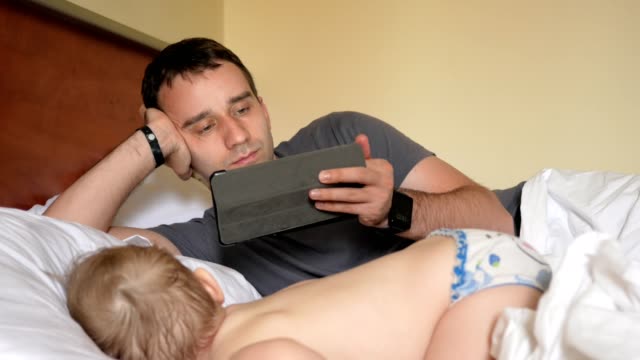 Vater-liest-die-Nachrichten-auf-dem-Tablett-in-der-Nähe-des-schlafenden-Babys.-Ein-Junge-in-den-Vordergrund-eines-Hauses-auf-einem-Bett-ruhen