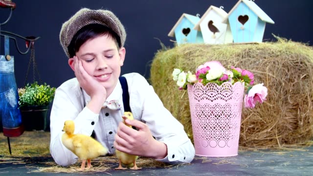 Dorf,-stilvoll-gekleidet-niedlichen-Jungen-spielen-mit-Enten-und-Hühner,-Studio-video-mit-thematischen-Dekor.-Im-Hintergrund-ein-Heuhaufen,-bunte-Vogelhäuschen-und-Blumen