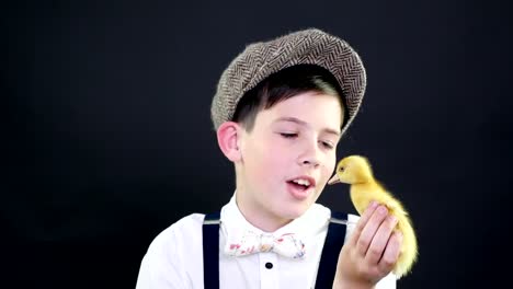 Portrait,-a-pretty-boy-in-a-cap-and-suspenders-plays-with-a-small-yellow-duckling.-Studio-video-with-thematic-decoration