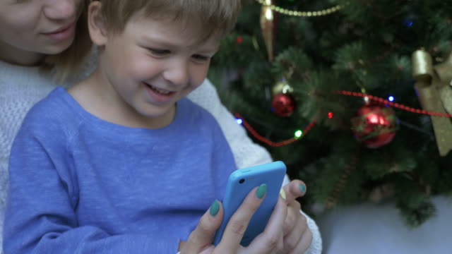 Fröhlicher-Junge-und-seine-Mutter,-Blick-auf-das-Smartphone-in-der-Nähe-von-Christmas-tree