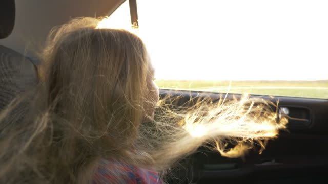 Young-caucasian-girl-looking-to-horizon-to-car-window.-Open-cars-window-and-strong-wind-fluttering-her-hairs.-Close-up-handheld-shot.-Traveling-by-car.-Autumn-sunset-backlit-light.-Sun-flare.