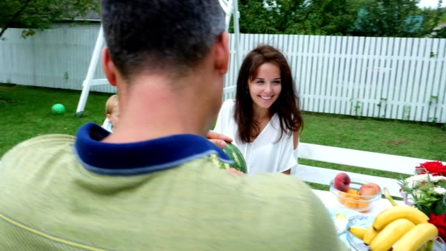 A-young-family-with-two-little-funny-children-sitting-at-a-dinner-table-in-the-garden,-in-summer.-Dad-cuts-a-large-watermelon,-is-going-to-treat-his-family.-lunch-with-the-family