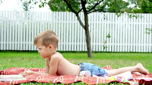 ein-vier-Jahre-alter-Junge-liest-ein-Buch-im-Garten,-im-Sommer-auf-einer-Decke,-einer-Tagesdecke,-Gras,-Rasen,-liegend