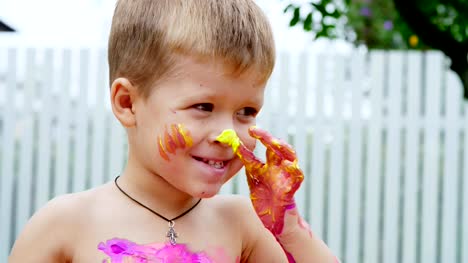 a-little-child,-a-four-year-old-boy-playing,-painting-with-finger-paints,-decorating-himself,-in-the-garden,-sitting-on-a-blanket,-on-grass,-lawn,-in-the-summer.-he's-having-fun