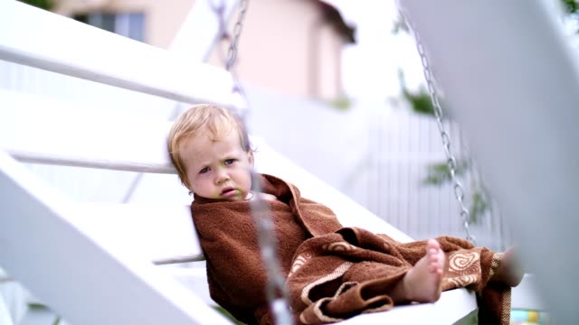 a-little-one-year-old-girl,-wrapped-in-a-towel,-with-a-dirty-face,-lonely-sitting-on-a-swing-in-the-garden,-in-the-summer.-She-has-a-sad-look.-She-wants-to-sleep