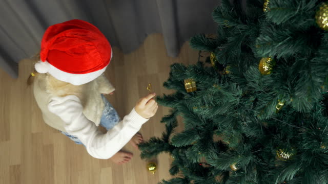 Happy-young-girl-in-red-Santa-hat-funny-and-happy.-White-girl-decorating-christmas-tree.-Gold-ball-fall-to-floor-near-christmas-tree.-Girl-repair-golden-sphere-decorative-toy.