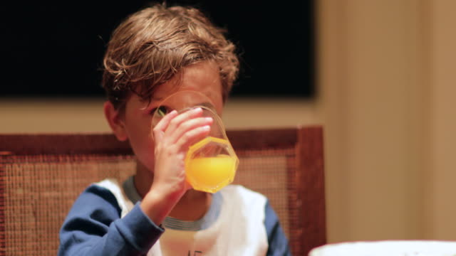 Niño-bebiendo-jugo-de-naranja.-Cándida-joven-tomando-un-sorbo-de-jugo-de-naranja-en-mesa