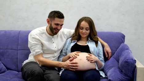 Pregnant-woman-sitting-at-home-on-the-couch-with-her-husband-watching-television.-A-man-communicates-with-his-wife-and-future-child.-Kiss-the-belly-and-listen-to-the-baby's-movements-in-the-belly