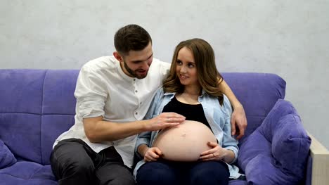 Pregnant-woman-sitting-at-home-on-the-couch-with-her-husband-watching-television.-A-man-communicates-with-his-wife-and-future-child.-Kiss-the-stomach-and-listen-to-the-baby's-movements-in-the-abdomen.-Hand-stroking-the-pregnant-belly.-Smile-and-laugh