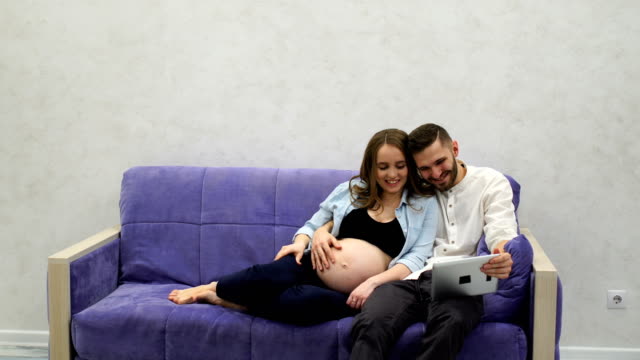 Happy-family-couple-sitting-on-the-couch-calling-on-video-communication-to-their-parents-in-another-city.-Tells-news-about-pregnancy.-A-pregnant-woman-and-a-man-communicate-via-a-tablet-computer-via-video-communication.-Video-conference.-Lifestyle-and