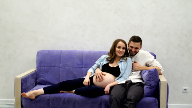 Glückliche-Familie-paar-auf-Sofa-vor-dem-Fernseher-sitzen.-Eine-schwangere-Frau-lacht-mit-ihrem-Ehemann,-während-gerade-eine-TV-show