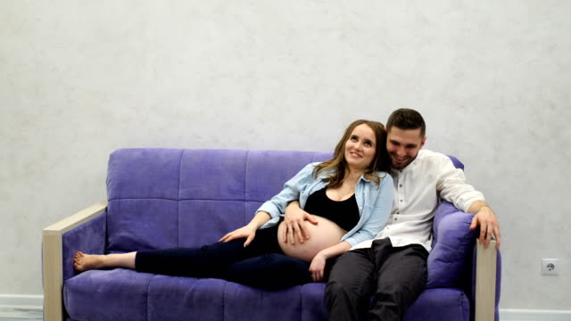 A-married-couple-is-sitting-at-home-on-the-couch-waiting-for-the-birth-of-a-child.-The-man-stroked-her-hand-across-her-belly-and-kissing-the-belly-of-his-pregnant-wife