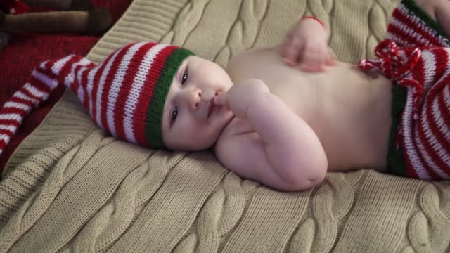 Friedliche-Babyjungen-gekleidet-in-gestreiften-Mütze-und-Hose-liegt-auf-Bett,-einen-Finger-saugen-und-lächelnd