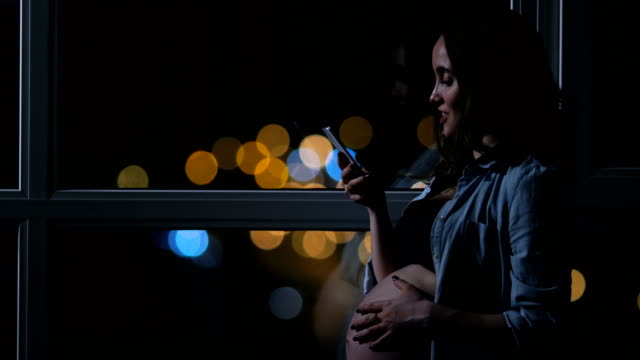 Pregnant-woman-at-night-talking-on-a-mobile-phone-touching-his-stomach-standing-at-the-large-panoramic-window-overlooking-the-city.-Cars-drive-by-outside-the-window.-The-girl-looks-at-the-phone-screen.-beautiful-bokeh-in-the-background