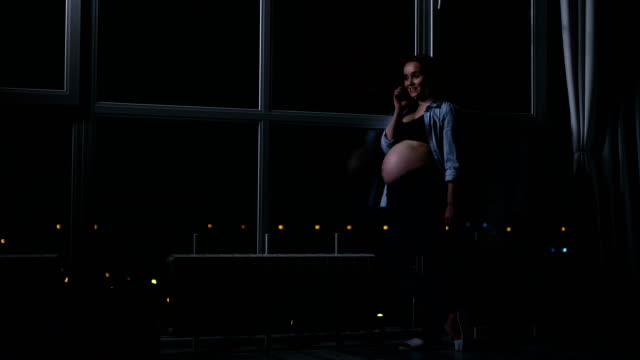 Una-mujer-embarazada-en-la-noche-hablando-en-un-teléfono-móvil,-tocando-su-estómago-con-las-manos-en-la-gran-ventana-panorámica-con-vistas-a-la-ciudad