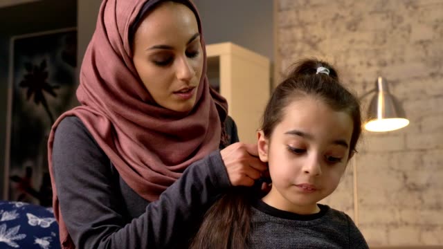 A-young-Indian-mother-wearing-a-hijab-makes-a-hairstyle-to-her-little-daughter-and-talks,-while-child-reading-a-book,-sitting-on-couch,-family,-home-cosiness-concept,-close-up-50-fps