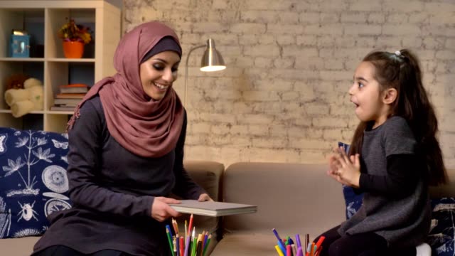 Young-Muslim-mother-in-a-hijab-takes-a-book-in-her-hands,-her-daughter-enthusiastically-laughs-and-claps-her-hands-50-fps