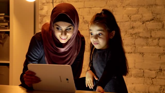 Una-joven-madre-en-un-hiyab-se-sienta-con-su-pequeña-hija-en-el-sofá-en-la-noche-y-utiliza-una-tableta,-un-vídeo-divertido,-riendo,-confort-en-el-hogar-en-el-fondo-50-fps