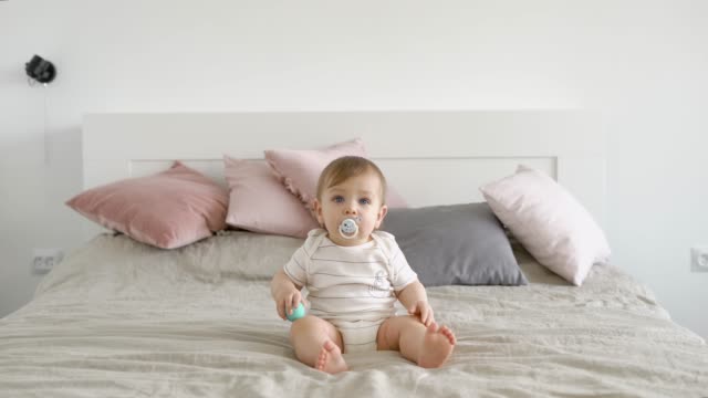 blonde-blue-eyed-infant-is-sitting-in-a-large-bed-with-baby-pacifier-and-looking-forward-in-daytime-in-cozy-home