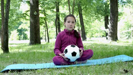 Happy-little-girl-sitting-in-the-city-Park-on-the-grass-in-a-pink-suit-holding-a-soccer-ball,-looking-at-the-camera-and-smiling.-4K,-29.97-fps.