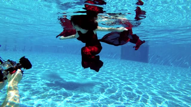 A-man-photographer-takes-a-professional-underwater-camera-beautiful-girl-bride-with-red-hair,-who-swims-underwater-in-the-pool-in-a-red-dress.-Slow-motion.