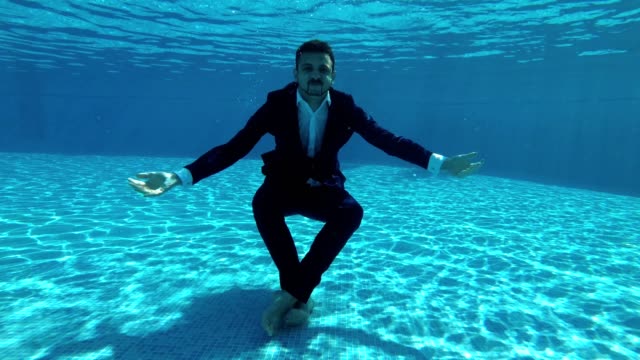 Asian-man-in-a-suit-and-a-white-shirt-dives-into-the-water-in-the-pool.-He-looks-at-the-camera-and-waves-his-hands.-Shooting-action-camera-under-water.