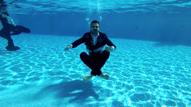 A-man-businessman-in-a-suit-and-a-white-shirt-dives-to-the-bottom-of-the-pool-under-the-water.-He-sits-on-the-bottom-in-a-Lotus-position,-looks-into-the-camera-and-lets-out-bubbles.-Slow-motion.-Action-camera-underwater.
