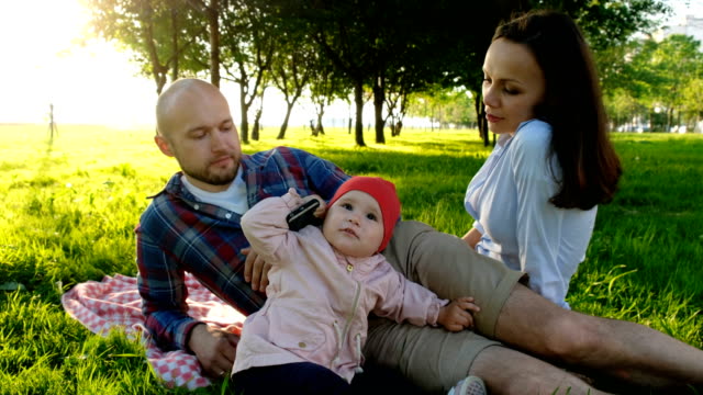 Bebé-niña-hablando-por-teléfono,-con-un-smartphone-en-sus-manos.-Familia-feliz-teniendo-un-picnic-al-aire-libre-en-verano-al-atardecer