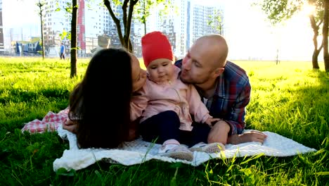 Happy-family-lies-on-grass-in-summer-park.-Parents-kiss-a-little-baby-girl-on-the-cheeks-on-both-sides