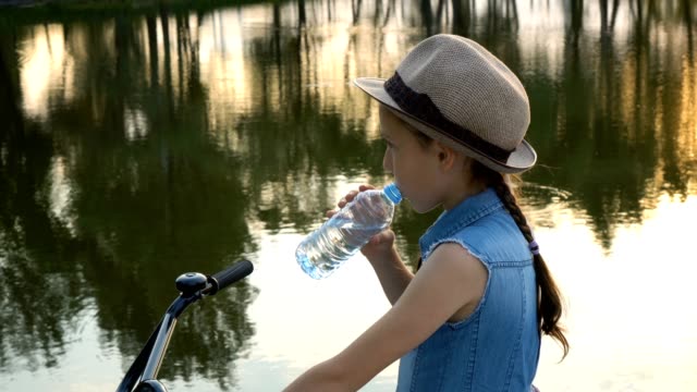 Happy-little-girl-with-a-bike-looking-at-the-camera-and-smiling-on-the-river-Bank-at-the-sunset.-She-stands-in-a-hat-on-a-background-of-water-and-drinks-water-from-a-clear-bottle.