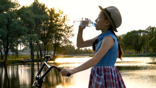 Hermosa-pequeña-niña-agua-potable-de-una-botella-transparente-al-atardecer.-Ella-está-con-un-sombrero-con-una-bicicleta-en-Banco-Río-en-la-luz-del-sol.
