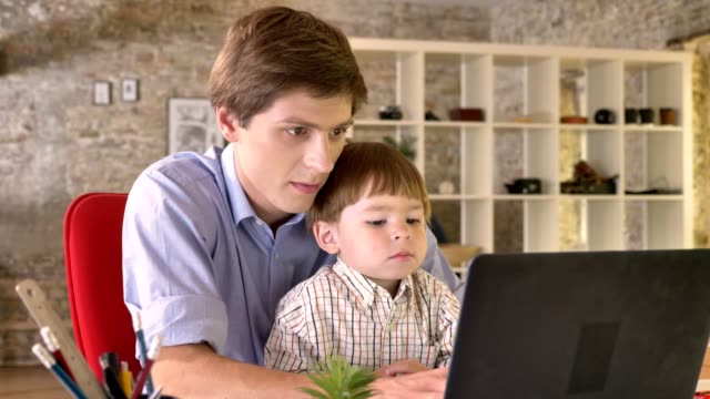 Joven-padre-sosteniendo-a-su-pequeño-hijo-y-trabajando-en-ordenador-portátil,-ocupado,-sentado-en-la-oficina-moderna
