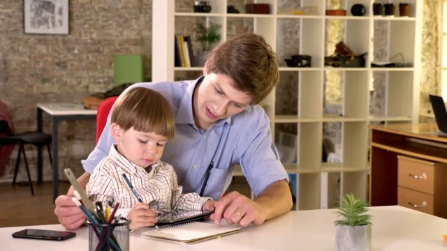 Junge-kaukasischen-Vater-hält-seinen-Sohn-und-ihm-beizubringen,-wie-man-schreiben,-sitzen-im-modernen-Büro
