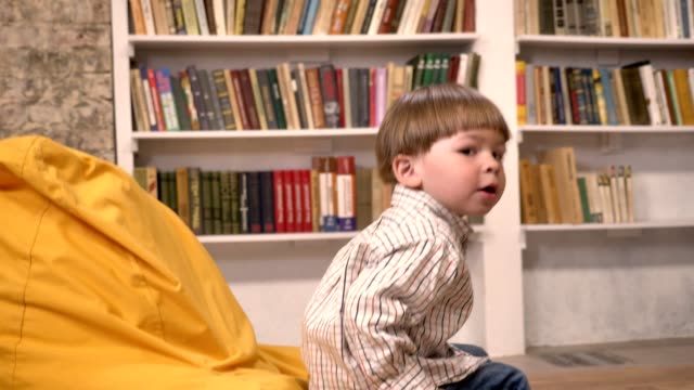Kleine-süße-Kind-auf-Stuhl-sitzend-und-Ball-von-jemandem,-Bücherregale-Hintergrund-fängt
