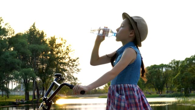 Sporty-little-girl-drinking-water-after-Cycling.-She-stands-on-the-background-of-the-river-at-sunset-and-drinks-from-a-transparent-plastic-bottle.-Bottom-view.-4K.-29.97-fps.