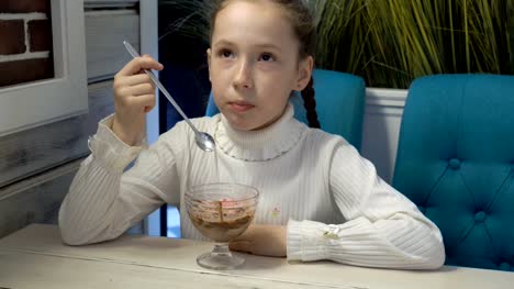 Linda-niña-con-coletas-sentada-y-comiendo-helado-de-fresa-y-chocolate-en-el-café.-Ella-come-con-una-cuchara-larga-de-cristal-taza.-Retrato