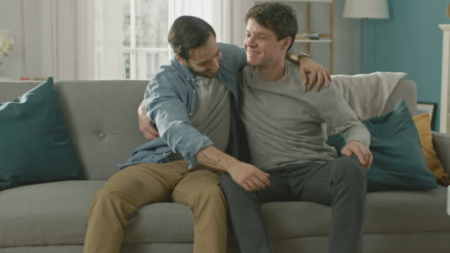Cute-Attractive-Male-Gay-Couple-Sit-Together-on-a-Sofa-at-Home.-Boyfriend-Puts-His-Hand-on-Partner's-and-They-Hug.-They-are-Happy-and-Smiling.-They-are-Casually-Dressed-and-Room-Has-Modern-Interior.
