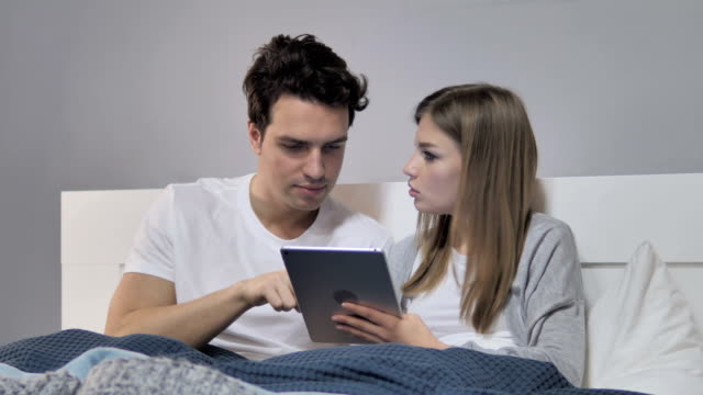 Relaxing-Young-Couple-Browsing-Internet-on-Tablet-in-Bed