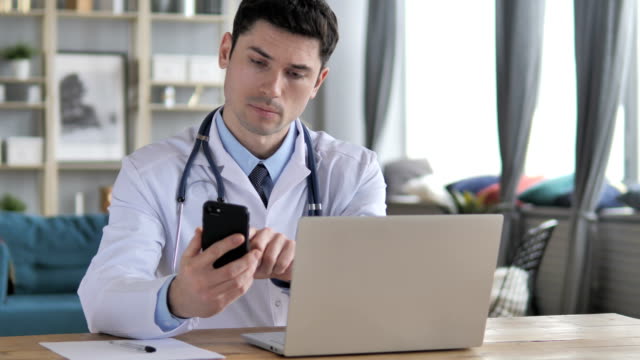 Arzt-am-Laptop-arbeiten-und-mit-Smartphone