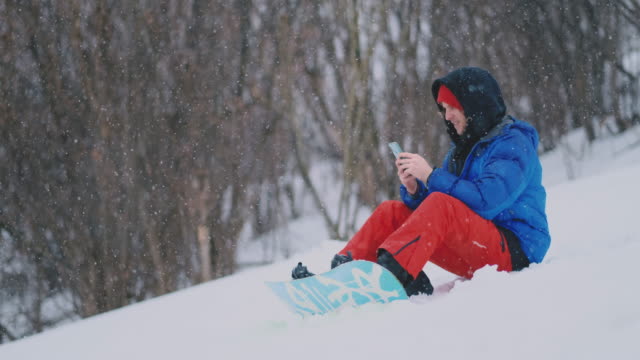 Ein-männlicher-Snowboarder,-der-auf-dem-Schnee-sitzt,-fotografiert-am-Telefon-eine-schöne-Resort-Landschaft-für-soziale-Netzwerke.-Blogger-im-Resort.-Schreiben-Sie-Textnachrichten-an-Freunde-mit-Ihrem-Smartphone