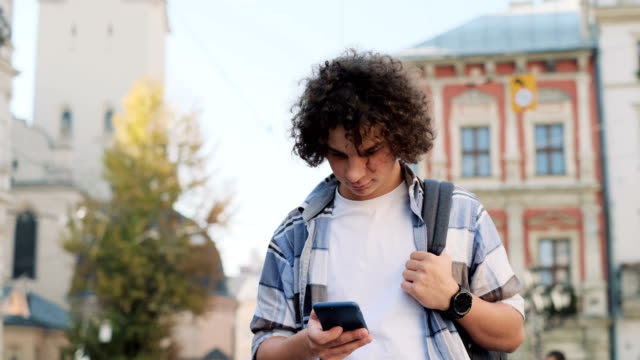 Young-curly-man,-tourist-or-student,-generation-z-in-hipster-outfit,-walking-through-the-street,-using-his-smartphone-scrolls-through-social-media-feed-on-device,-checking-map-or-reading-news-on-app