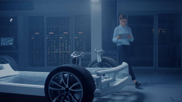 Automobil-Ingenieur-arbeitet-auf-Elektro-Auto-Chassis-Plattform,-mit-Tablet-Computer-Augmented-Reality-mit-3D-CAD-Software-Modellierung.-Innovative-Anlage:-Fahrzeugrahmen-mit-Rädern,-Motor,-Batterie