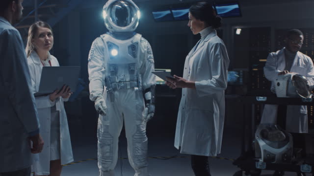 Team-von-Luft-und-Raumfahrtingenieuren-Design-New-Space-Suit-für-Galaxy-Exploration-und-Reisen-angepasst.-Gruppe-von-Wissenschaftlern-Wearing-White-Coats-haben-Diskussion,-Verwendung-von-Computern.-Astronautenhelm-bauen