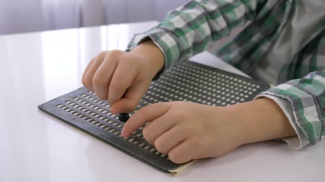 Niño-con-discapacidades-visuales-aprendiendo-a-escribir-caracteres-Braille-de-fuente-sentado-en-la-mesa-en-la-habitación-luminosa,-las-manos