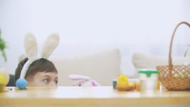 Cute-little-boy-with-bunny-ears-is-hiding-under-the-table-full-of-Easter-decorations.-Little-cute-white-bunny-is-attacking-boy-kindly.-Laught-is-in-the-room.