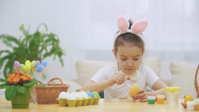 Little-playful-girl,-wearing-bunny-ears-on-her-head-is-choosing-an-a-red-colour-to-paint-an-egg-and-calmly-is-painting-an-Easter-egg.