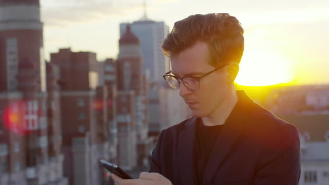Stylischer-Mann-mit-Smartphone-auf-Rooftop-bei-Sonnenuntergang